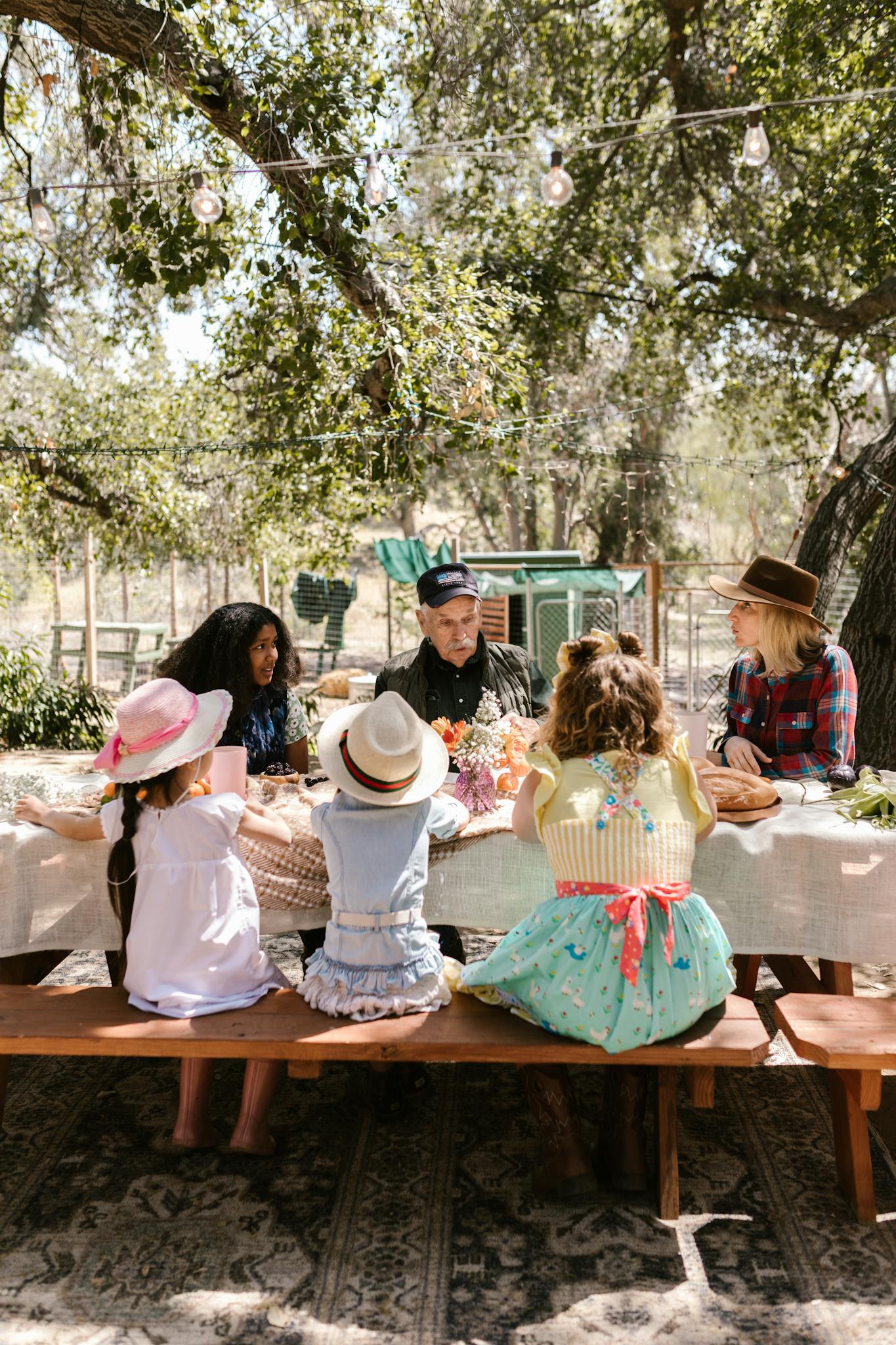 A family enjoying a gathering at an outdoor farm table under trees.